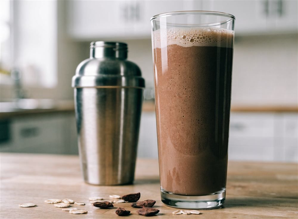 A glass of chocolate protein shake for kids next to a shaker bottle on a kitchen counter